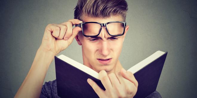 A young man lifting his glasses straining to read a book.